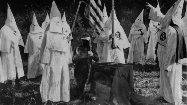Iniciação de novo membro da Ku Klux Klan - 10 pessoas com vestes da Ku Klux Klan e um homem de terno sentado, rosto coberto, mão para cima em juramento