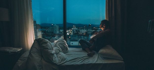 Man listening to music on headphones at dusk, looking through the window while sitting on his bed.