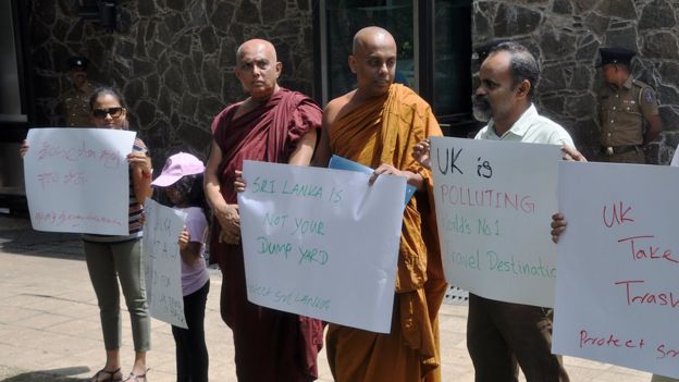 A small crowd of people hold hand-written protest signs outside a guarded building in this photograph