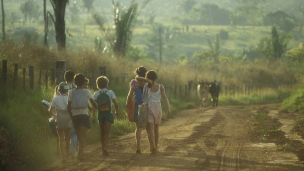 Crianças com uniformes e mochilas andam em estrada de terra em cena rural