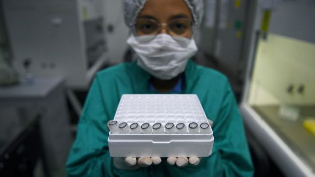 Funcionária de máscara e jaleco mostra caixa com amostras a serem testadas para covid-19