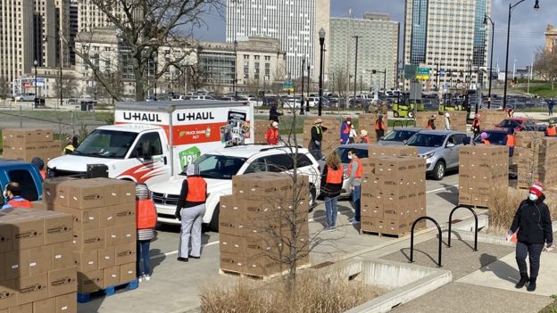 Fila de carros recebendo caixas com alimentos doados entregues por voluntários usando máscaras e coletes em cidade dos Estados Unidos