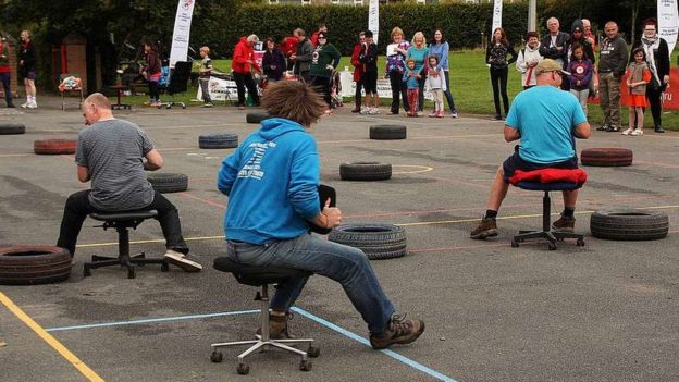 Three men race around a course on office chairs watched by a crowd