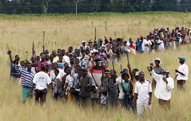 Veteranos de guerra em 2000 marcham em um campo, segurando enxadas