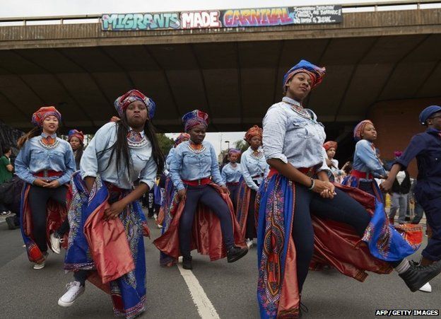 Performers dance in the street beneath a sign reading Migrants Made Carnival