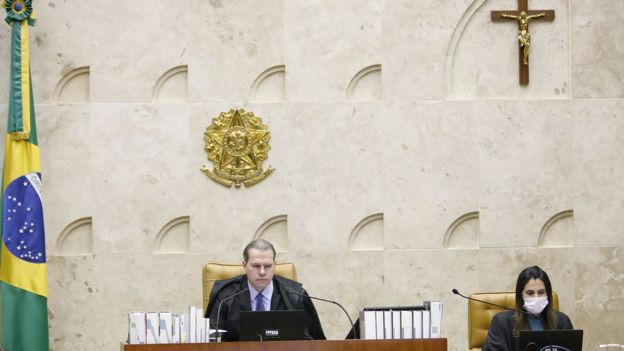 Dias Toffoli sentado no centro do plenário do STF, ao lado de assistente com máscara