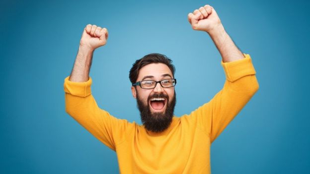 A smiling, young, bearded man in eyeglasses and yellow shirt holding hands up in feeling of happiness and gratitude - blue background.