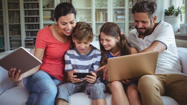 Stock photo of a family of four sitting on a sofa, using a laptop, mobile and a tablet