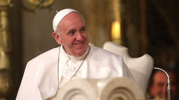 O Papa Francisco, com as vestimentas papais, senta em uma cadeira e sorri