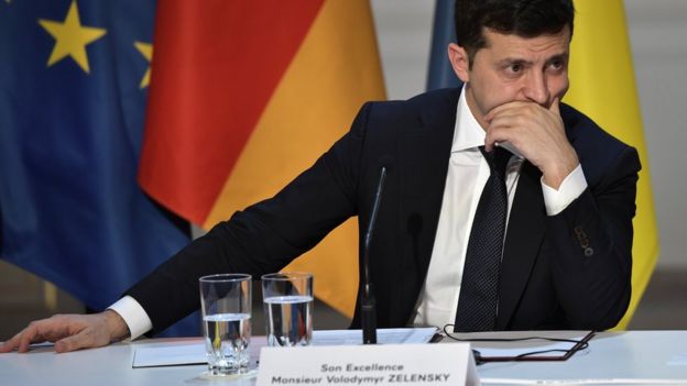 Volodymyr Zelensky aparece sentado em mesa de evento, com bandeiras atr&aacute;s