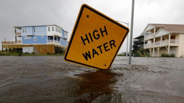 Imagem mostra placa informando que o nÃ­vel da água está alto, em meio a enchente e casas na Carolina do Norte