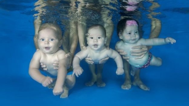 Três bebês debaixo d'água, segurados por adultos