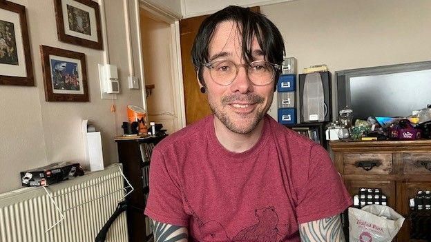 A man with dark brown hair. He is sat down in a room in a house and wearing a red t-shirt.