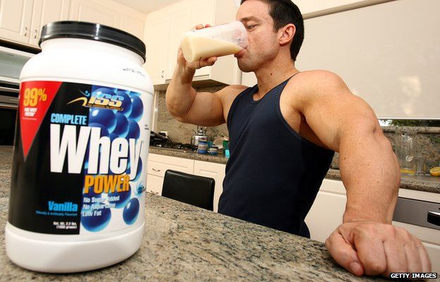A large tub of whey protein powder sits on a kitchen worktop as a bodybuilder takes a drink