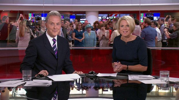 Julian Worricker and Sophie Long present the news as Queen Elizabeth II is shown around the crowded BBC newsroom behind them