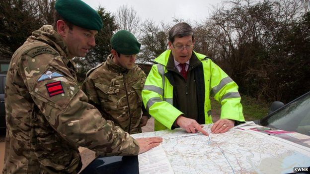 Two soldiers and a man in a high-visibility yellow jacket looking at a map placed on the bonnet of a car