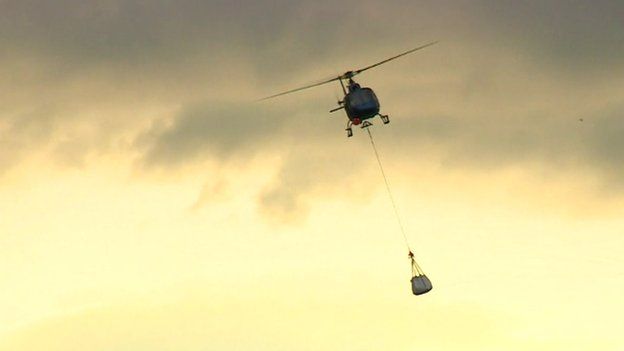 A helicopter carrying a large bag of slate suspended beneath it by a rope