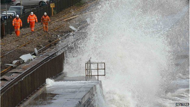 A wave crashes onto a damaged section of railway track, as repair workers walk nearby