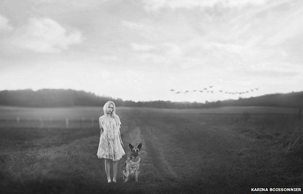 A girl stands barefoot in a field with a German shepherd dog. In the distance a flock of birds flies in formation.