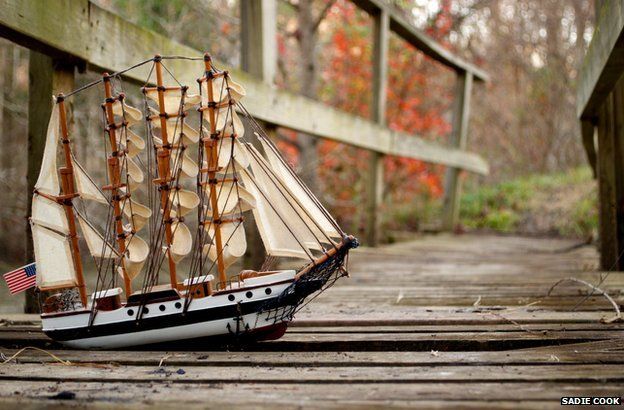 A model of an old-fashioned sailing vessel with full sails and a US flag, is pictured on a bridge in a rural setting.