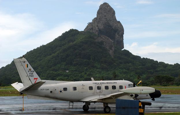 A small Brazilian Air Force radar plane prepares to leave an airport strip in front of a green, rocky, hill