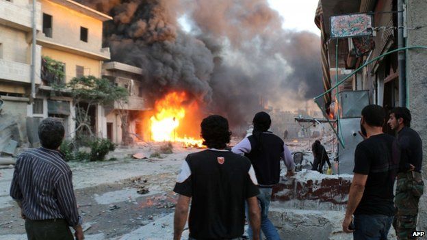 Rebel fighters and civilians stand looking at a burning building following a reported barrel bomb attack by Syrian government forces in Aleppo, 20 April 2014