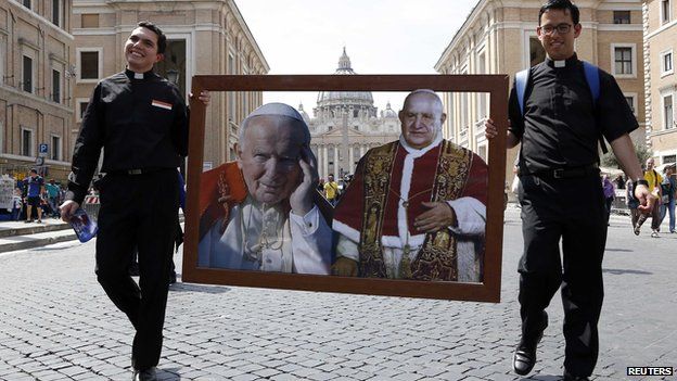 Two priests walk with pictures of Pope John Paul II (L) and Pope John XXIII in St Peter's square, Rome, on 25 April 2014.