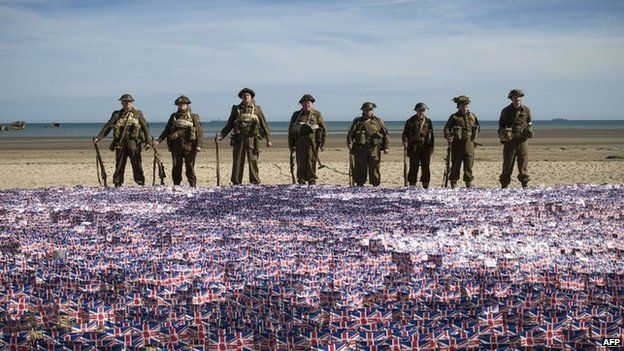 People in WW2 uniforms standing behind many small union flags on a beach