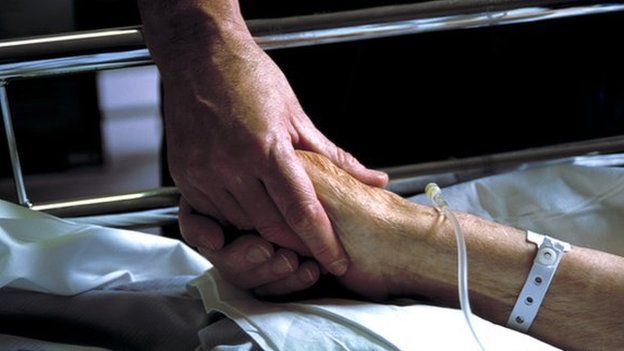 Two people holding hands. One is lying in a hospital bed with an intravenous drip.