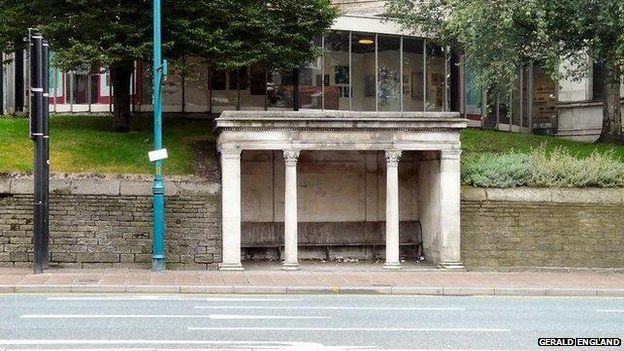 A stone shelter with wooden benches on Wellington Road South. Behind it is Stockport Art Gallery.