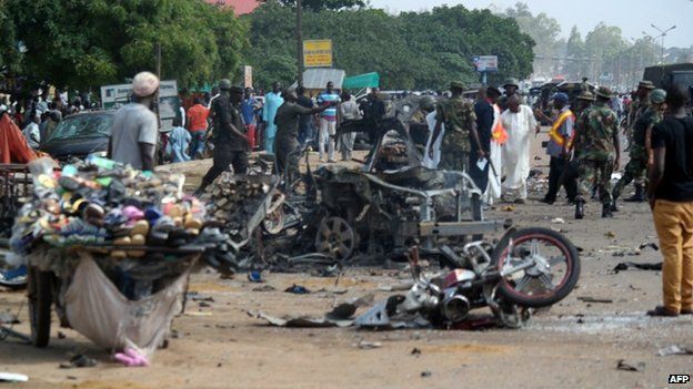 People stand near a wrecked car after a bomb exploded on July 23, 2014 in Kaduna, north of Nigeria
