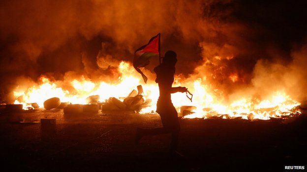 A Palestinian protester carried a flag past burning tyres at Qalandia, on the West Bank, 24 July 2014