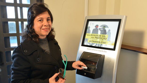 Professor Liliae Ventura stands in front of a kisok-like machine holding a pair of sunglasses
