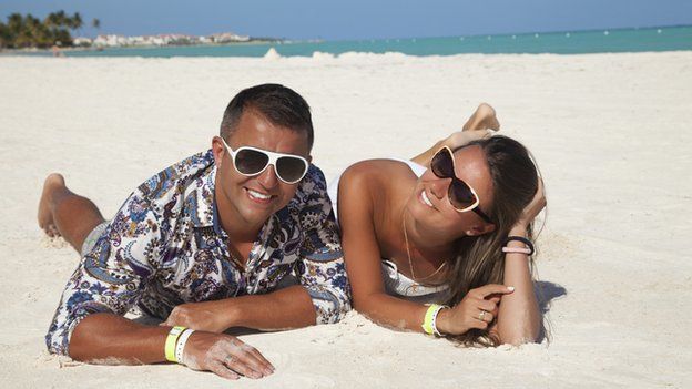 A man and a woman, both wearing sunglasses, lie on a sandy beach looking at the camera. The sea is in the distance behind them.