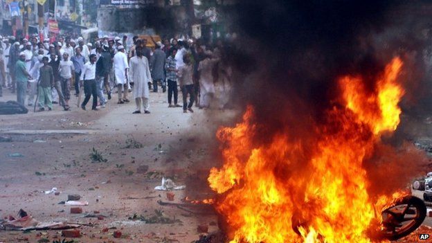 Indians stand near a burning motorcycle, set on fire during a protest between two communities in Saharanpur district of Uttar Pradesh state, India, Saturday, July 26, 2014