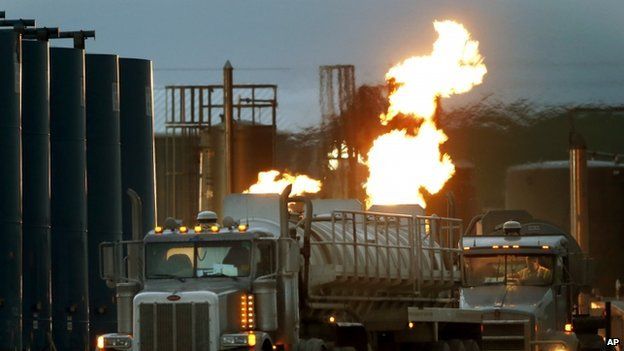 Tanker trucks, capable of hauling water and fracking liquid line up near a natural gas burn off flame and storage tanks.