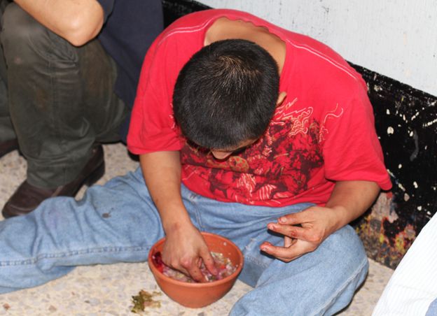 A man sits on the floor against the wall, eating out of a bowl