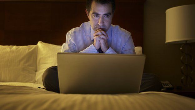 A man in a hotel room looks at a laptop.