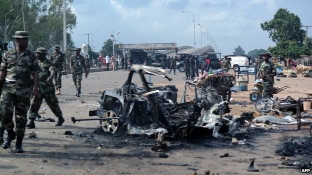 Military officers walk past the remains of a car after an explosion on 23 July 2014 in Kaduna, north of Nigeria.