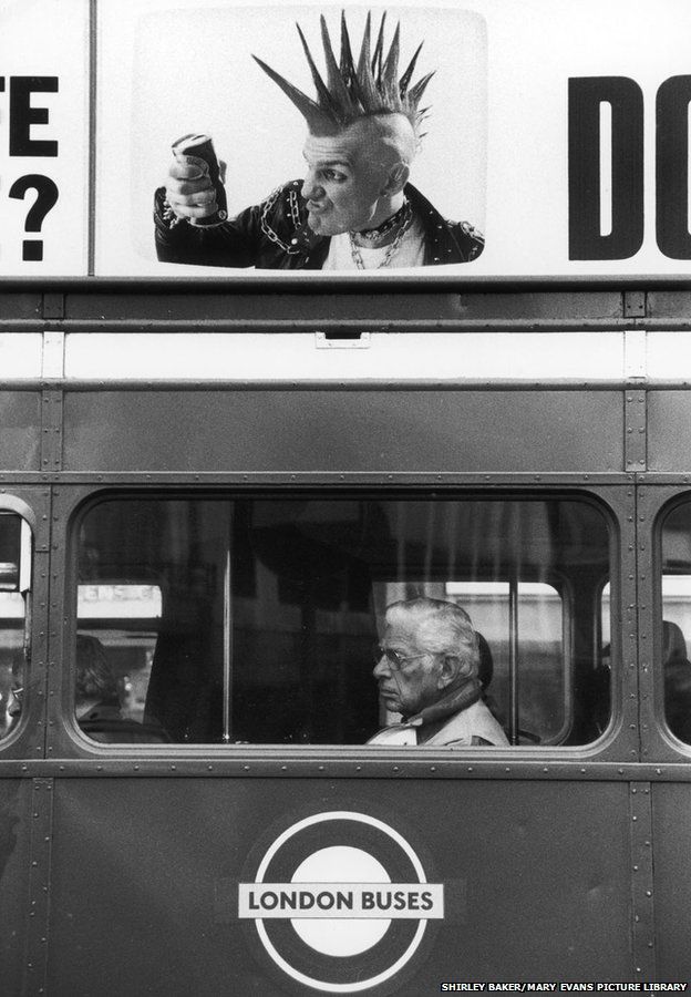 A terrific photograph showing the striking juxtaposition of a neat if stern elderly bus passenger framed in the window of the bus, set below an advert on the side of the vehicle depicting an angry punk