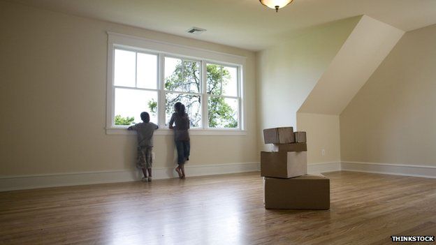 Two children stare out of a window in an empty room with boxes