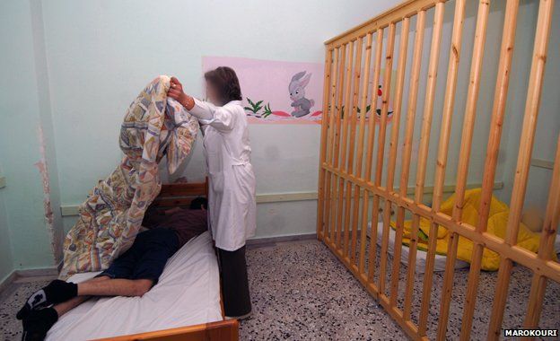 Wooden bars separate part of a room - a person lies behind the bars and a woman puts a cover over someone lying on a bed, 2008