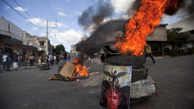 Image of Haiti's Prime Minister Laurent Lamothe stands against burning tyres in Port-au-Prince. 5 Dec 2014