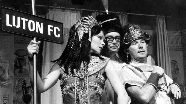 Glenda Jackson is dressed up as Cleopatra holding a Luton FC sign, with Eric Morecambe as Octavian Caesar and Ernie Wise as Mark Anthony