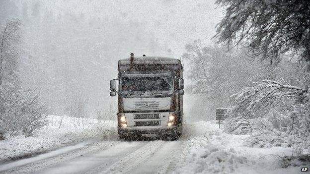 A lorry driving along a snow-covered road, as snow continues to fall