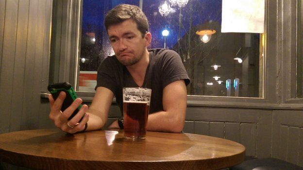 William in the pub holding his phone looking morosely at it