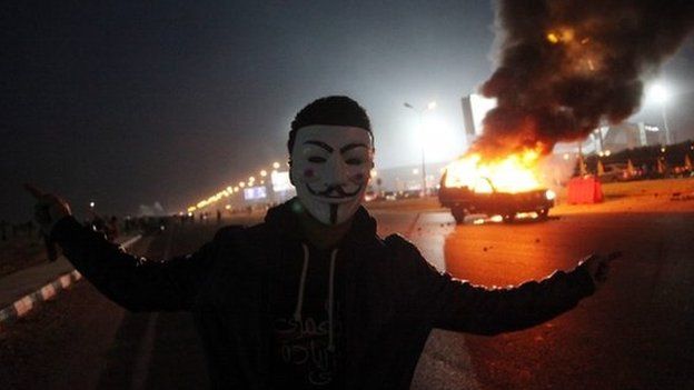 An Egyptian Zamalek soccer club fan wearing a Guy Fawkes mask near a burning police car outside a sports stadium in Cairo"s northeast district