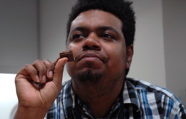 Ishmael Buckner holds a piece of chocolate, preparing to taste it. He has a thoughtful look on his face.