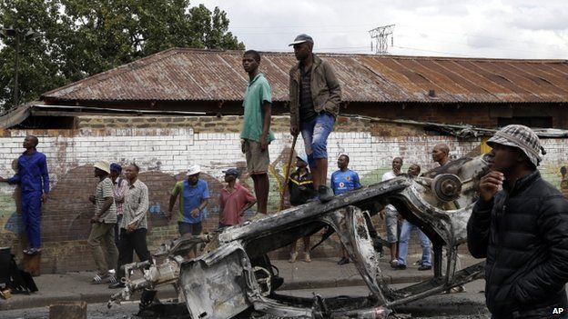 A crowd of anti-immigrant protesters stand on a burned-out car outside the Jeppe hostel in Johannesburg, South Africa, Friday, April 17, 2015
