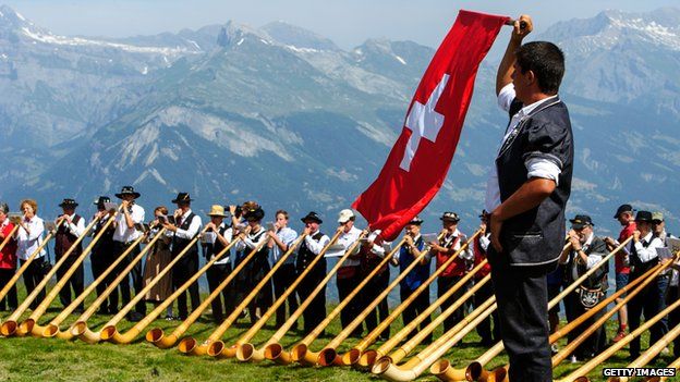 A man throws a Swiss flag as alphorn players perform on July 28, 2013 in Nendaz, Switzerland. About 150 alphorn blowers performed together on the last day of the international Alphorn Festival of Nendaz. The Swiss folkloric wooden wind instrument was used in most mountainous regions of Europe by mountain dwellers as signal
